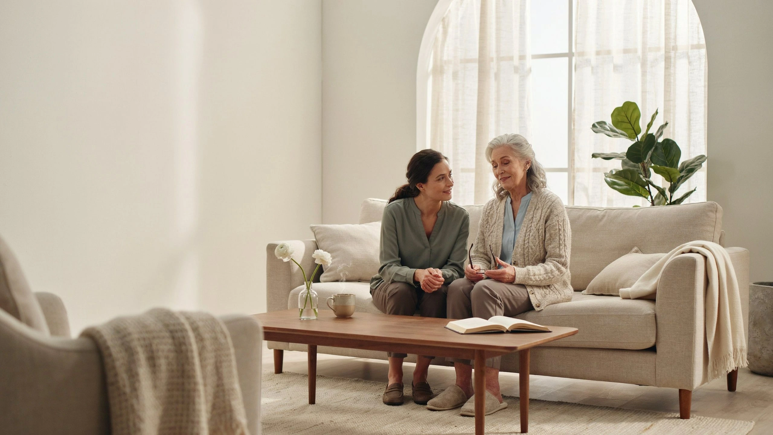Caregiver sitting beside a senior woman on a couch, sharing a relaxed conversation in a cozy living room