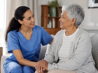 Smiling caregiver in blue scrubs holding hands with a senior woman on a couch at home