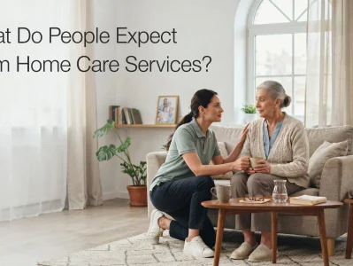 A caregiver in a light green polo shirt kneels beside an elderly woman on a sofa, placing a hand gently on her shoulder while handing her a cup, with text reading "What Do People Expect From Home Care Services?"